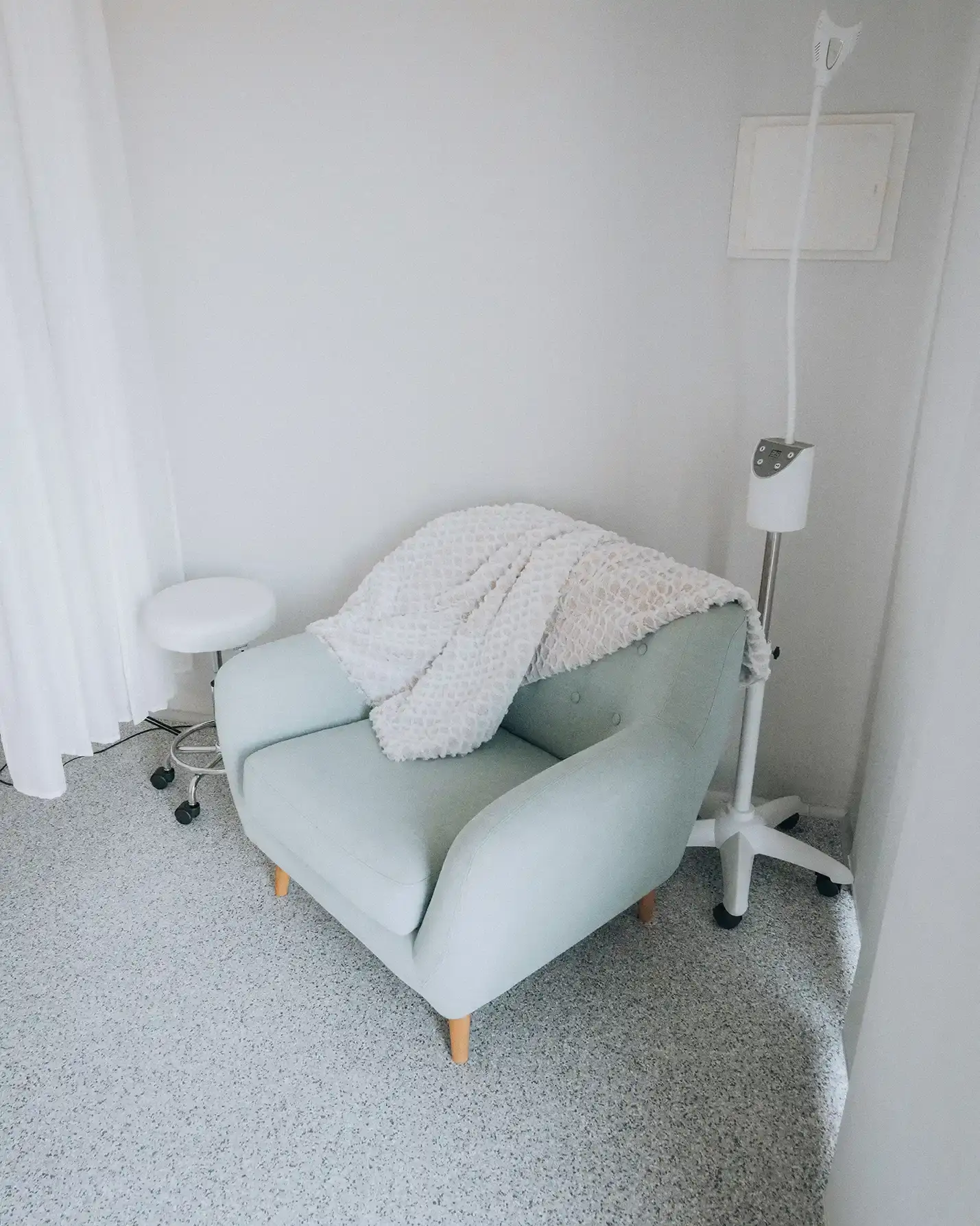 A sage green armchair with a soft throw over the back, beside a treatment lamp and stool, in the consultation corner at Enrich Aesthetics in Wangi Wangi