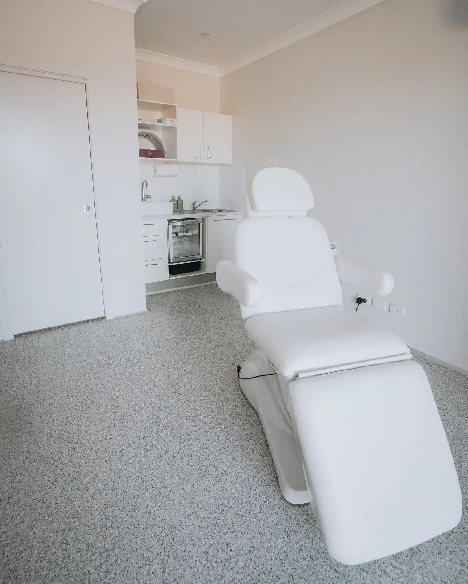 The reclining white treatment chair, with the clinic basin and fridge behind it